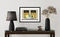 Framed artwork of a Prada store window on a wall above a table with decor items.