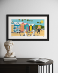 Framed artwork of beach huts and people on a wall above a table with a statue and book.