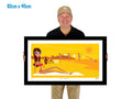 Man holding a framed artwork of a woman sunbaking on Bondi Beach with lifesavers hanging washing in background