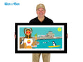Man holding a framed artwork of a woman at a beach with penguins and a building in the background.