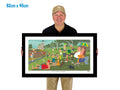 Man holding a framed artwork of a park scene with people and dogs.