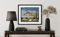 Framed artwork of a building on a table with a lamp and vase.