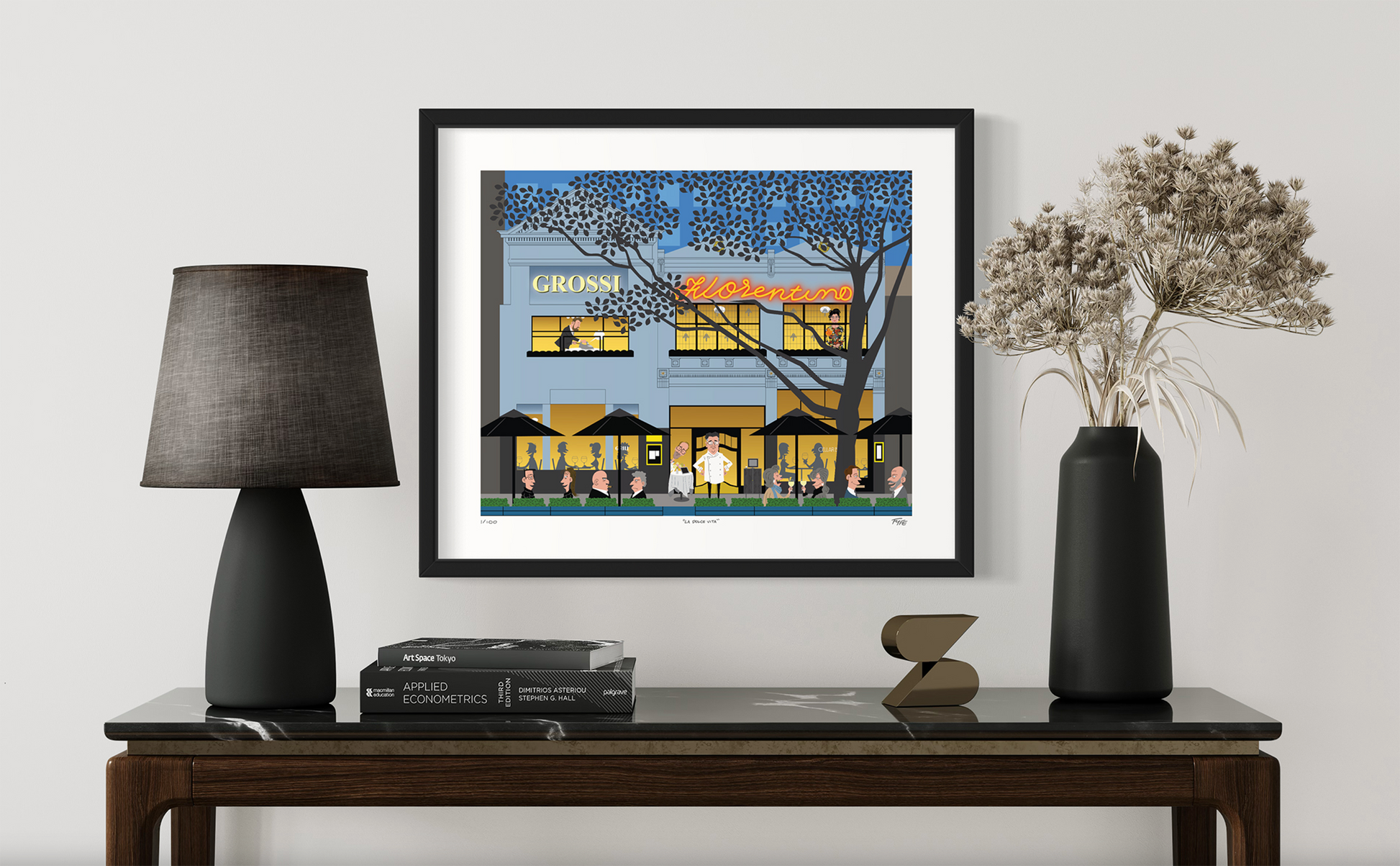 Framed artwork of a building on a table with a lamp and vase.
